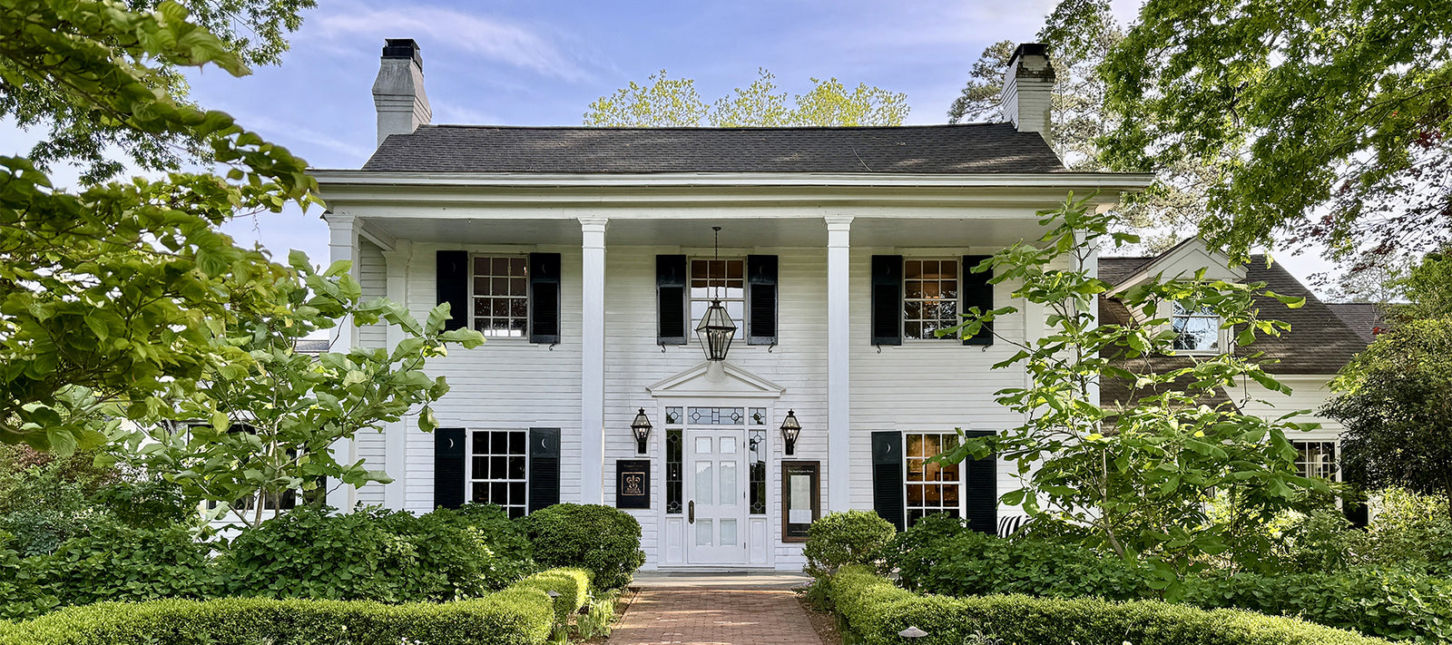A white two-story colonial-style house with black shutters, two chimneys, and four large front columns. Green shrubs and trees frame a brick pathway leading to the front door.