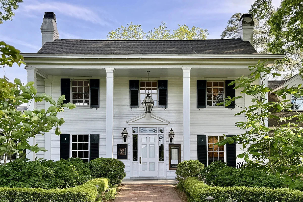 A white two-story house with black shutters, four columns at the entrance, and a central front door with sidelights. The house is surrounded by green shrubs and trees under a blue sky.