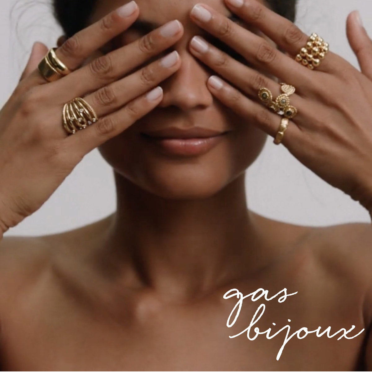 A woman with a slight smile covers her eyes with her hands, showcasing several ornate gold rings. The words 
