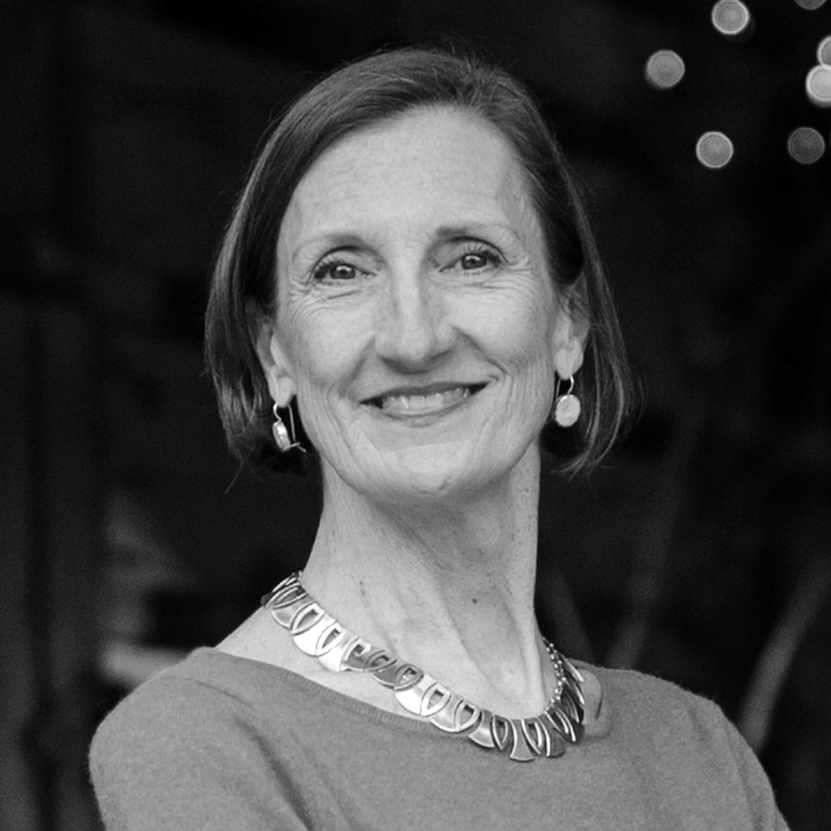 A woman with short hair smiles at the camera. She is wearing a necklace, earrings, and a top with a wide neckline. The photo is in black and white, with a blurred background and some circular lights visible.