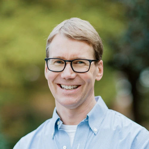 A smiling man with short light brown hair and glasses wearing a light blue button-up shirt, standing outdoors with greenery blurred in the background.