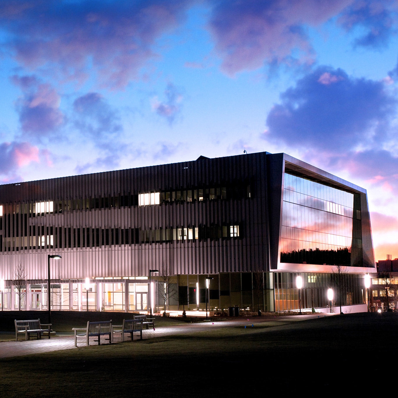 A modern, glass-walled building is illuminated at dusk with a colorful sunset reflecting in its windows. Benches and lights are visible on the grassy area in front of the structure.