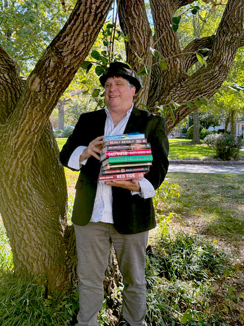 A smiling man in a black hat and blazer stands outdoors by a large tree, holding a colorful stack of books in his arms. Sunlight filters through the leaves, and a grassy yard is visible in the background.