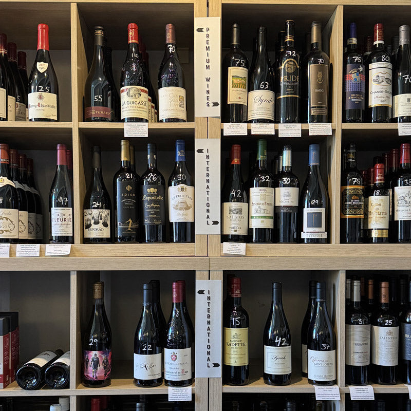 Rows of assorted wine bottles are displayed on wooden shelves. Labels, prices, and numbered tags are visible on each bottle. Signs indicate sections for "Premium Wines" and "International Wines.