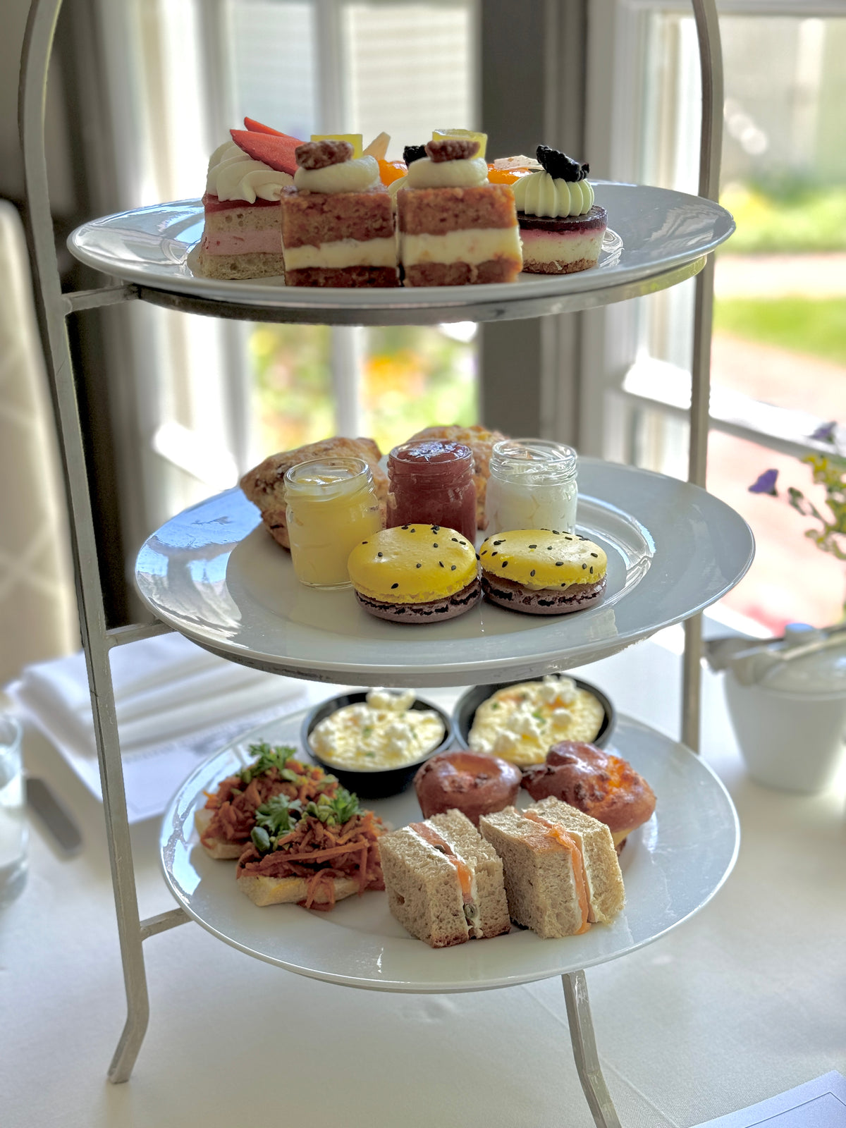 The Mother's Day Weekend Afternoon Tea from FEARRINGTON VILLAGE, which is now sold out, features a delightful three-tier cake stand. The top tier boasts an assortment of small cakes and tarts, the middle tier offers delicious scones accompanied by clotted cream and jam, while the bottom tier presents an array of finger sandwiches and pastries. This exquisite setup is beautifully arranged by a window at Fearrington House Restaurant.