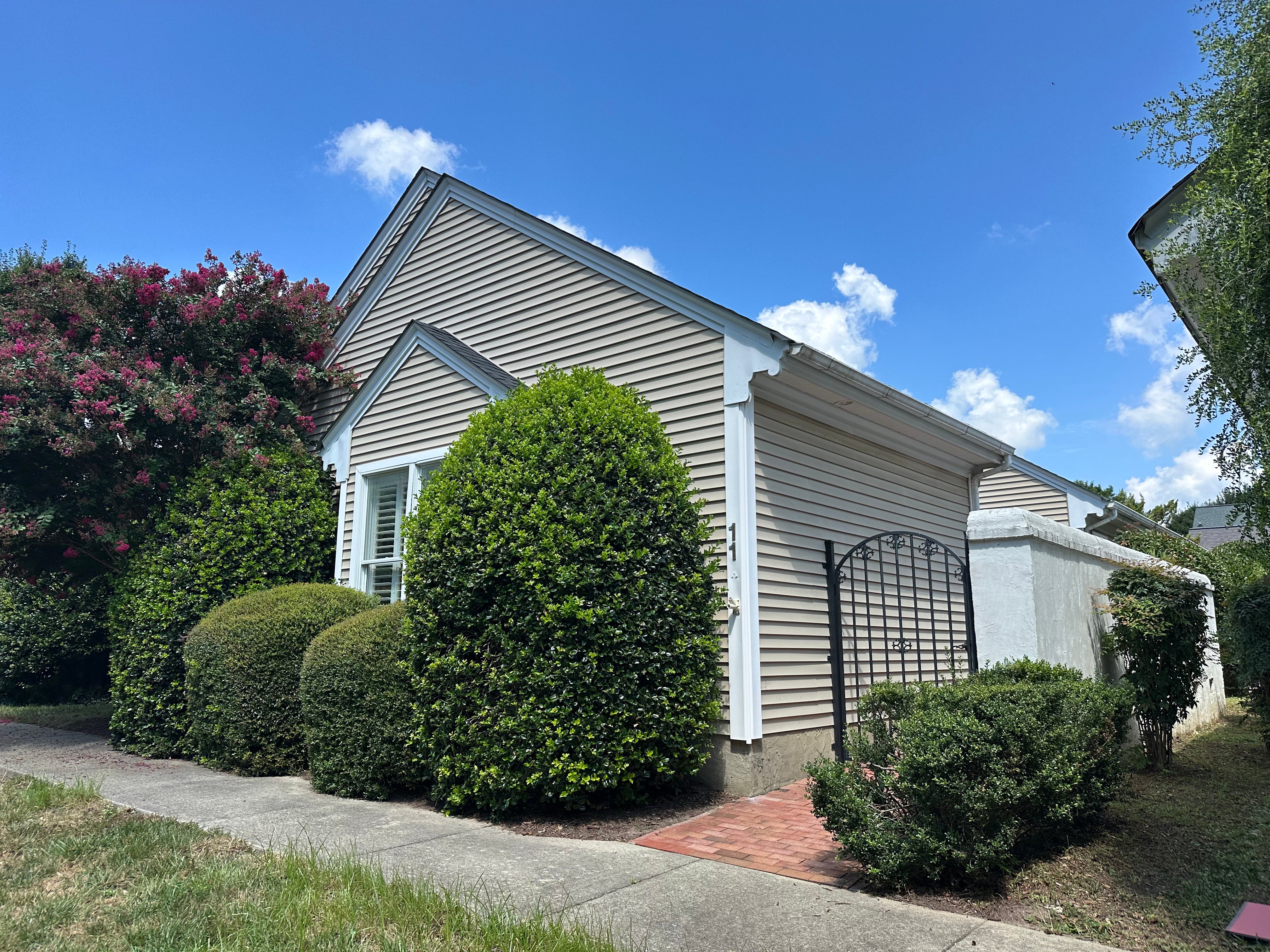 A small beige house with white trim, surrounded by neatly trimmed hedges and bushes. A black metal gate and brick pathway lead to the side yard. Blue sky with a few clouds is visible in the background.