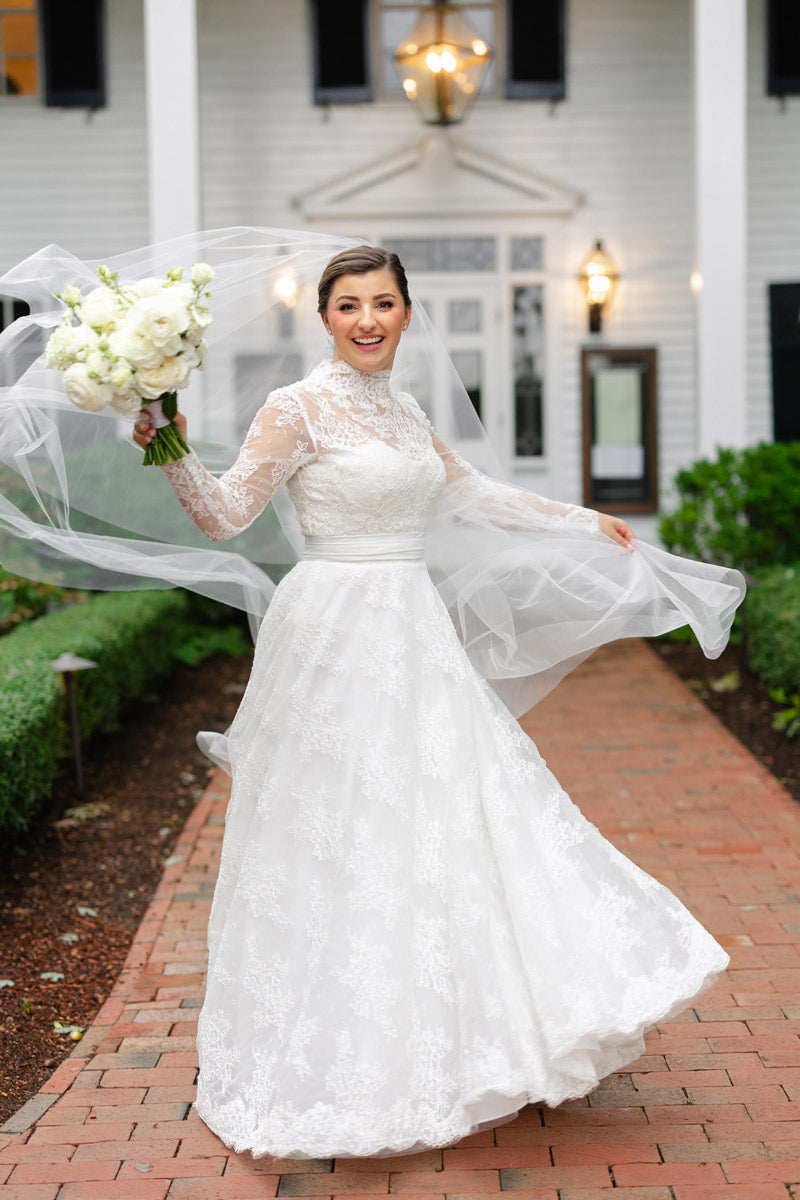 A smiling bride in a long-sleeved lace wedding dress holds a bouquet of white flowers and lifts her flowing veil, standing on a brick path in front of a white building with black shutters.