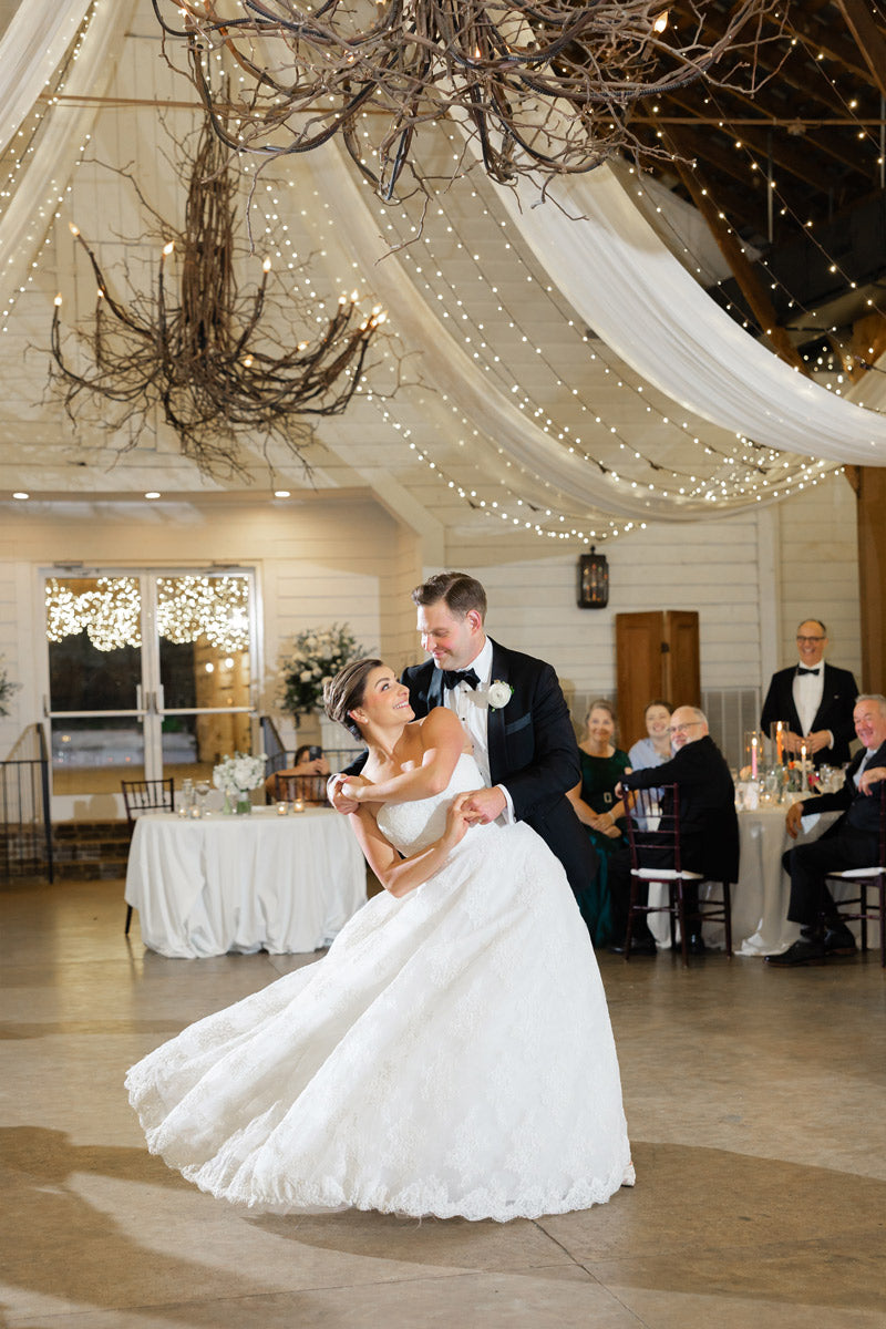 A bride and groom share their first dance in an elegant, rustic venue with draped fabric, string lights, and large branch chandeliers. Guests watch smiling from tables, while the bride laughs joyfully in a white gown.