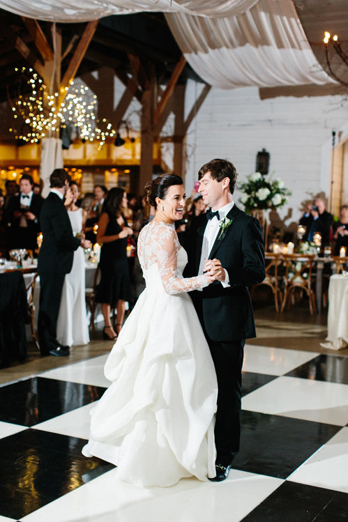 A bride in a long-sleeve white gown and a groom in a black tuxedo share their first dance on a black and white checkered floor at a wedding reception, surrounded by guests and elegant decorations.