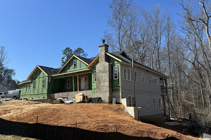 A house under construction on a dirt lot, with green sheathing on one side and finished gray siding on the other; surrounded by bare trees under a clear blue sky.