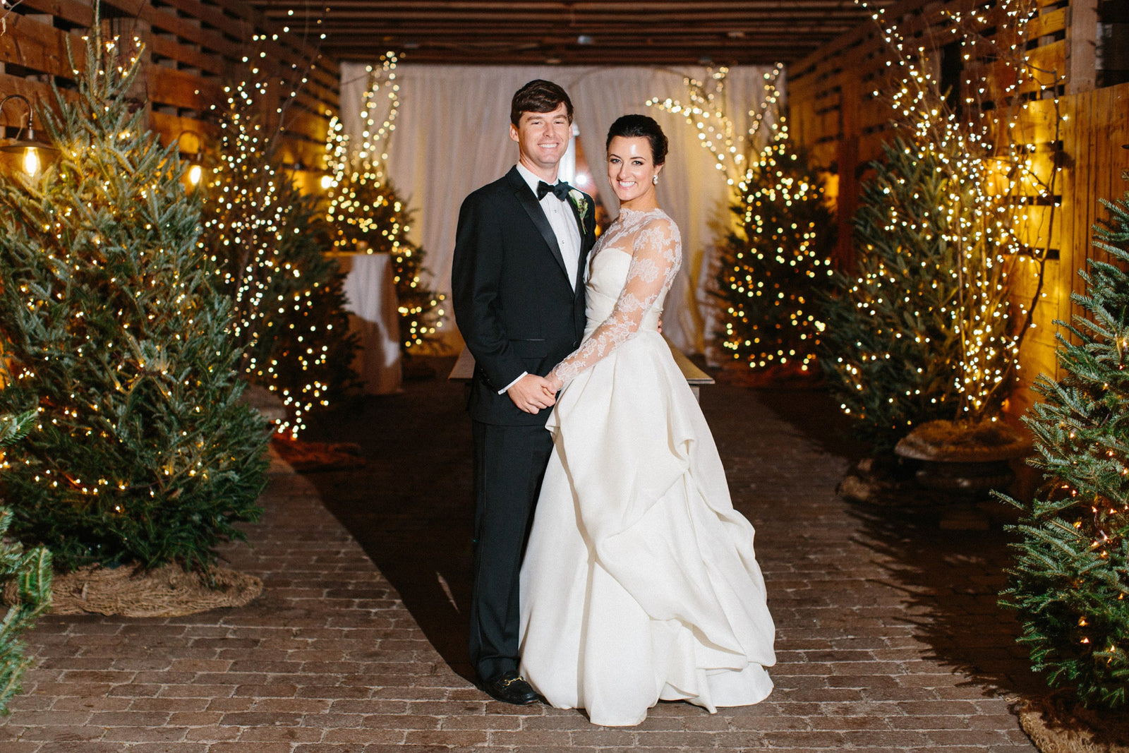 Man and woman in formal attire standing in a decorated indoor setting with Christmas trees.