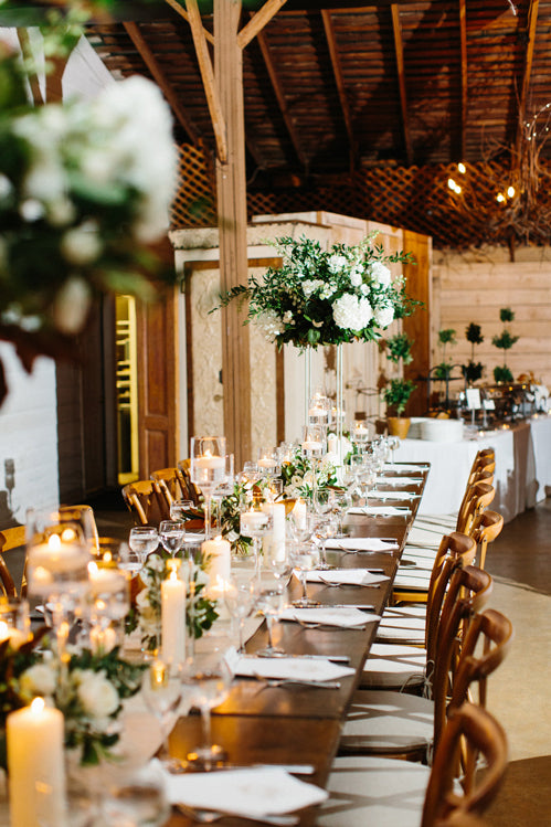 A long, elegant wooden table is set for a formal event with white flowers, green foliage, tall centerpieces, candles, glassware, and napkins in a warmly lit rustic venue with wooden beams.