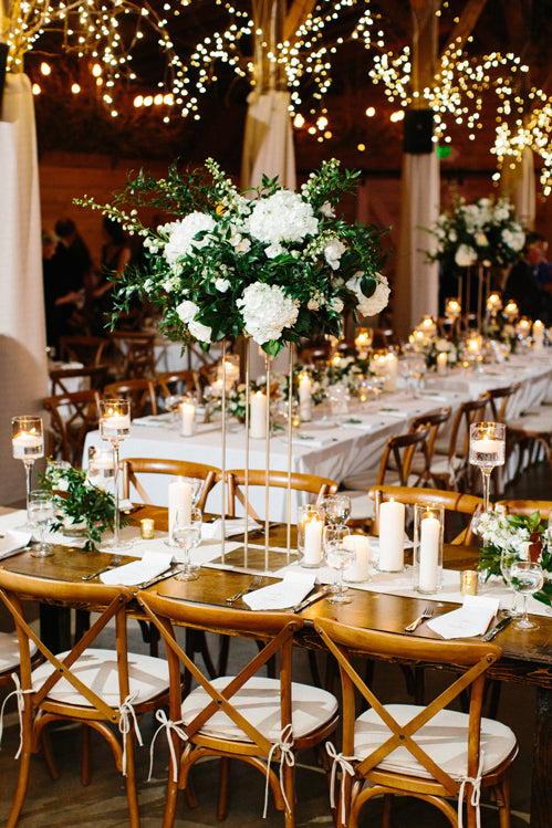 A long banquet table set for an event, decorated with tall floral centerpieces of white flowers and greenery, surrounded by wooden chairs and illuminated by string lights and candles.
