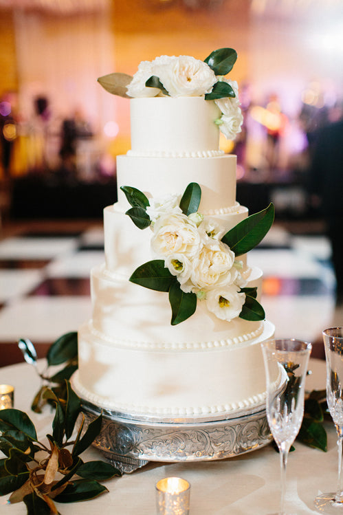 A four-tiered white wedding cake decorated with white flowers and green leaves sits on a silver stand, surrounded by champagne flutes and small candles on a table.