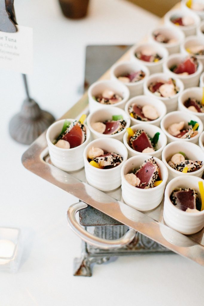 Small white cups filled with colorful appetizers, including slices of seared tuna, sesame seeds, greens, and a creamy topping, neatly arranged on a metal serving tray.