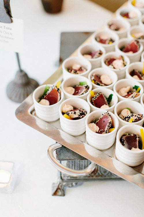 A tray of small white cups, each filled with colorful bite-sized appetizers, including pieces of seared tuna, greens, and sesame seeds, arranged neatly for a catered event.