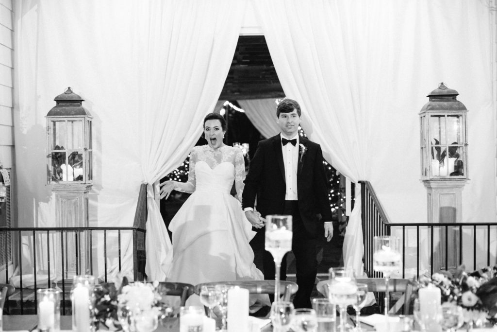 A bride and groom enter a decorated venue holding hands, both smiling excitedly. The bride wears a white dress and the groom a black tuxedo. Candles and flowers decorate the foreground. The scene feels festive and elegant.