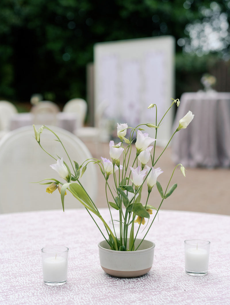 A small ceramic bowl with delicate white flowers and green stems sits on a round table covered with a light patterned cloth, flanked by two white votive candles, with blurred outdoor seating in the background.