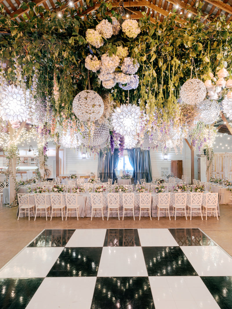 A lavishly decorated event hall with a black-and-white checkered dance floor, long tables set for dining, and floral arrangements and greenery hanging from the ceiling, creating a whimsical, garden-like atmosphere.