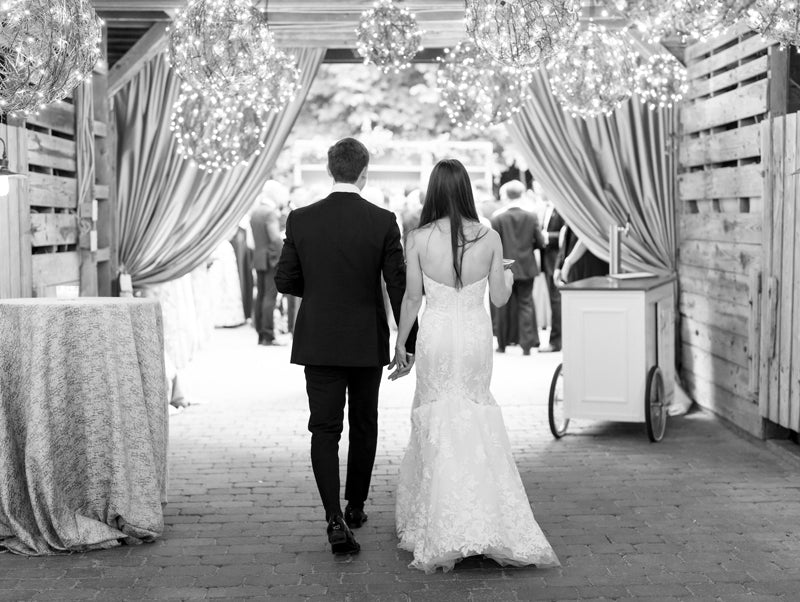 A bride and groom, seen from behind, walk hand-in-hand toward a group of people at an elegant indoor event space decorated with string lights and draped curtains.