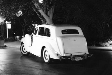 A vintage white car with classic design and round headlights is parked on a street at night, illuminated by streetlights and surrounded by trees.