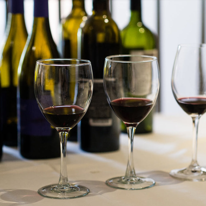 Three wine glasses with red wine rest on a white surface, bottles behind them—a scene from FEARRINGTON VILLAGE’s Wine Class: Blind Tasting 101, perfect for anyone seeking wine education or attending a sommelier class.