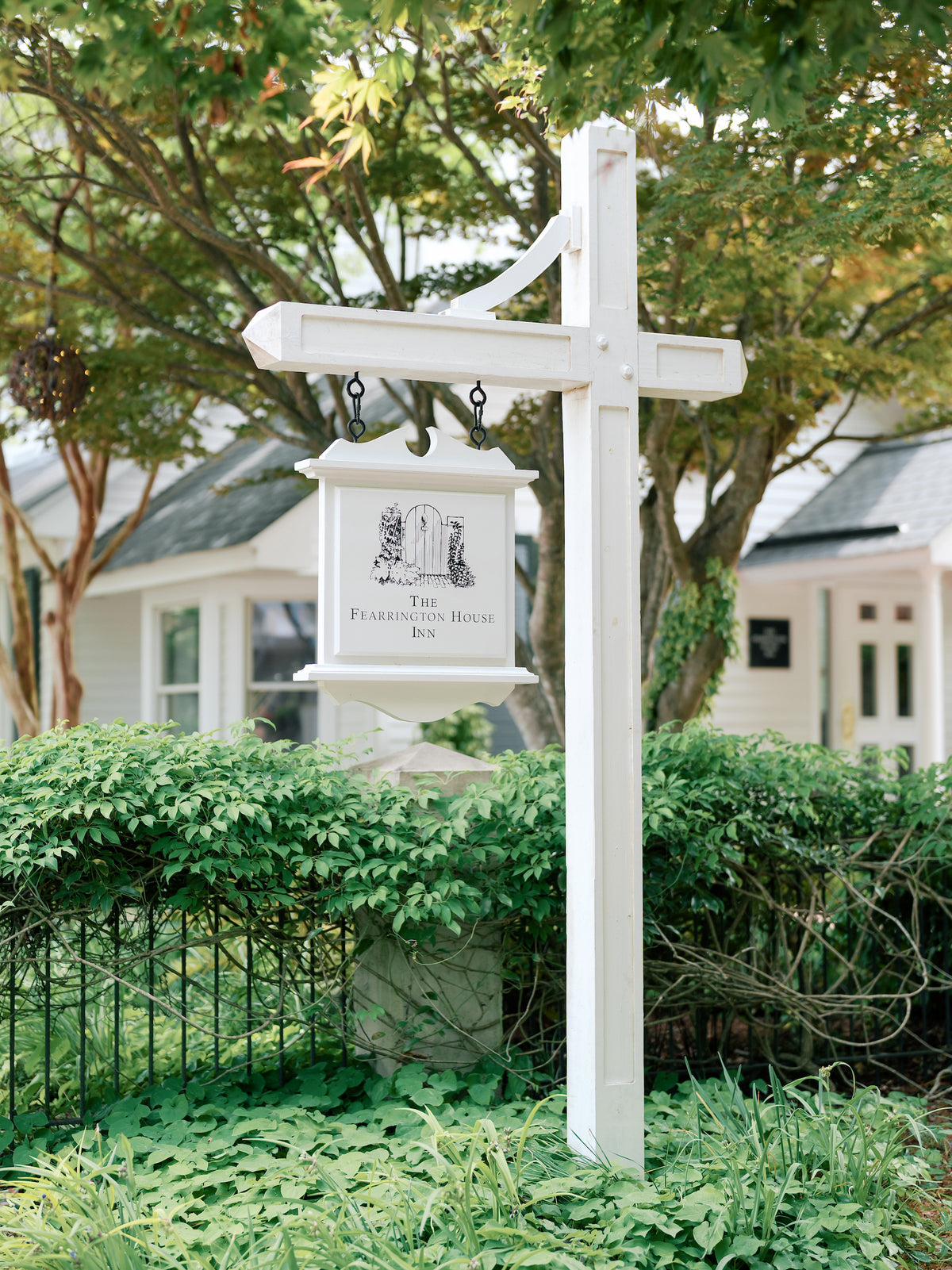 A white hanging sign reading "The Fearrington House Inn," with an illustration of a house, is mounted on a white post amid green foliage, in front of a white building and trees.
