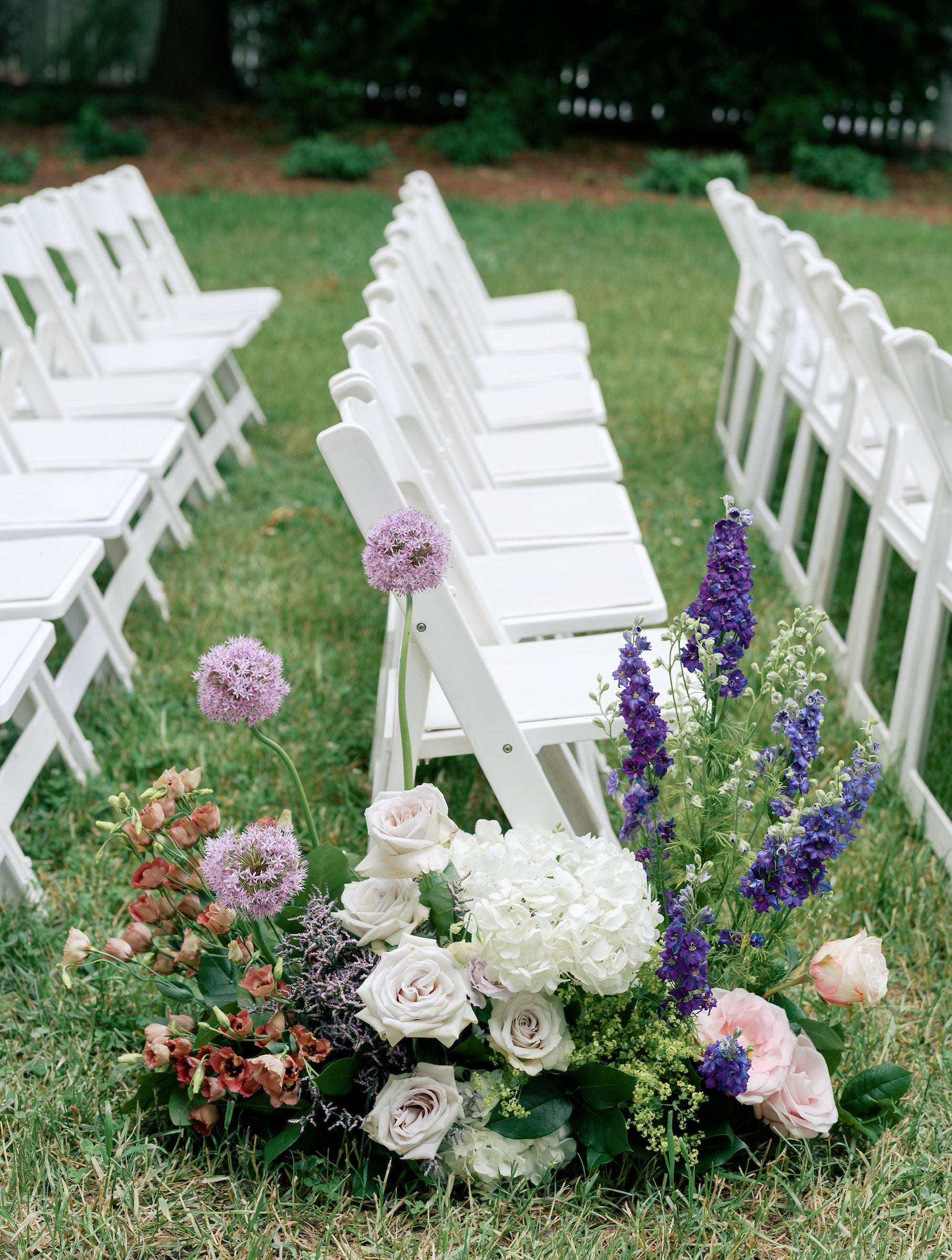 Rows of white folding chairs are arranged outdoors on green grass, with a floral arrangement of white, pink, and purple flowers placed at the end of one row, likely for a wedding ceremony.