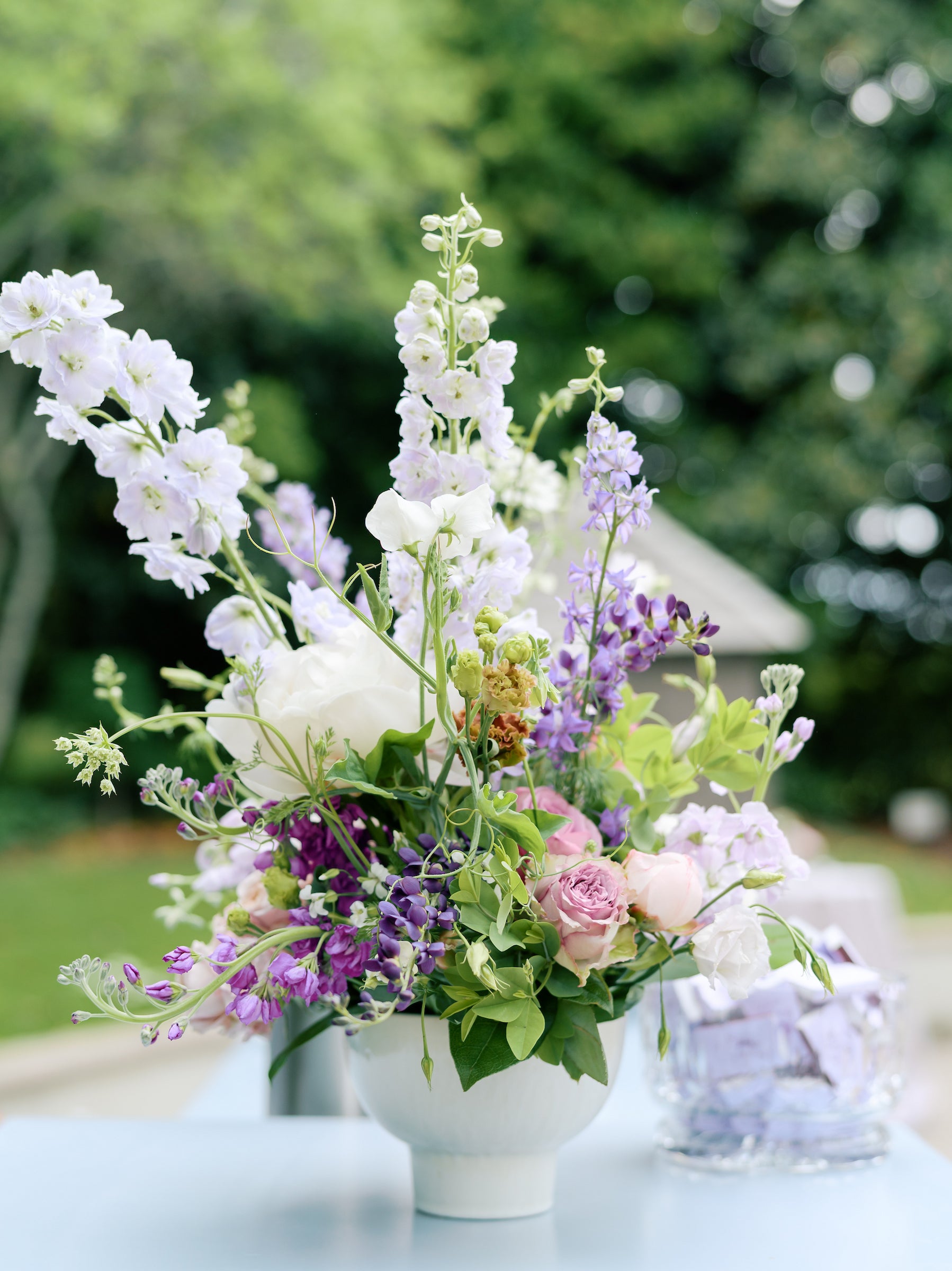 A white ceramic vase filled with a lush arrangement of pastel flowers, including white, purple, and pink blooms, sits on a light blue table outdoors with green trees in the background.
