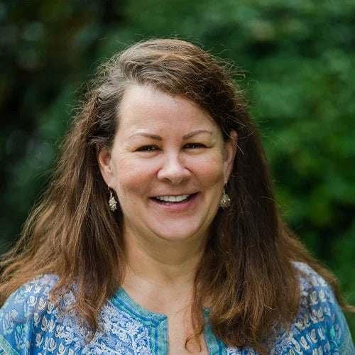 A woman with long brown hair, wearing a blue patterned top and dangling earrings, smiles outdoors with green foliage blurred in the background.