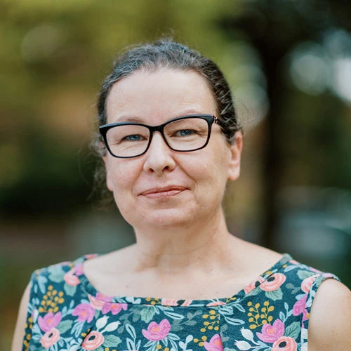 A middle-aged woman with glasses and pulled-back hair smiles softly, wearing a floral patterned top, standing outdoors with a blurred background of greenery.