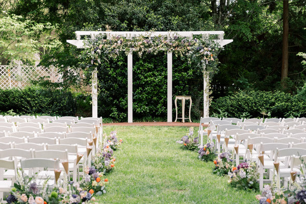 A white pergola decorated with flowers stands at the end of a grassy aisle lined with white chairs and floral arrangements, set outdoors among green trees and bushes, ready for a wedding ceremony.