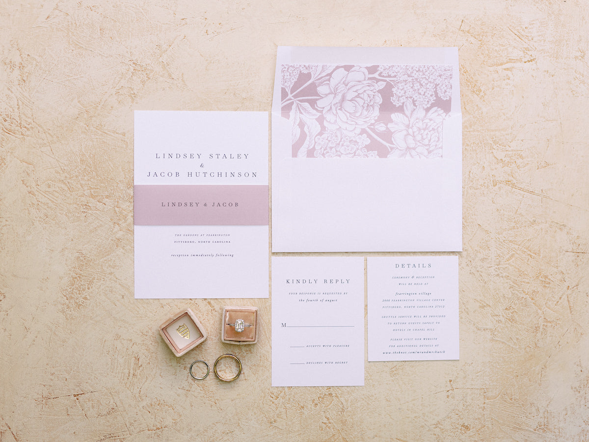 Wedding invitation suite displayed on a textured beige surface, including an invitation, RSVP card, details card, envelope with floral design, two rings, a ring box, and a wedding band. Colors are soft pink, white, and gold.