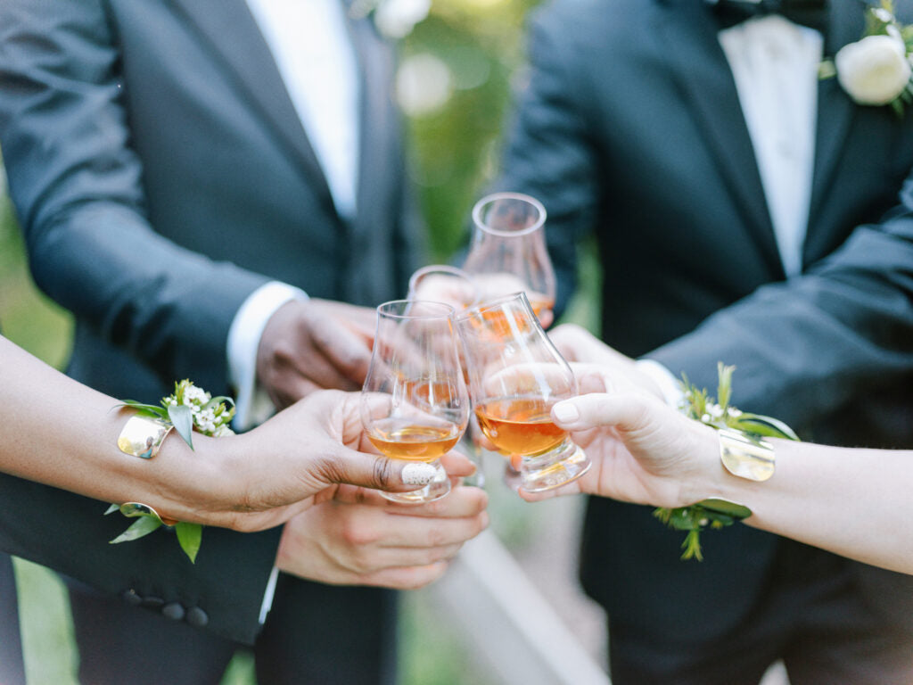 Four people dressed in formal attire clink glasses of amber-colored drinks in a celebratory toast. Each wears a boutonniere or corsage. The background is softly blurred greenery.