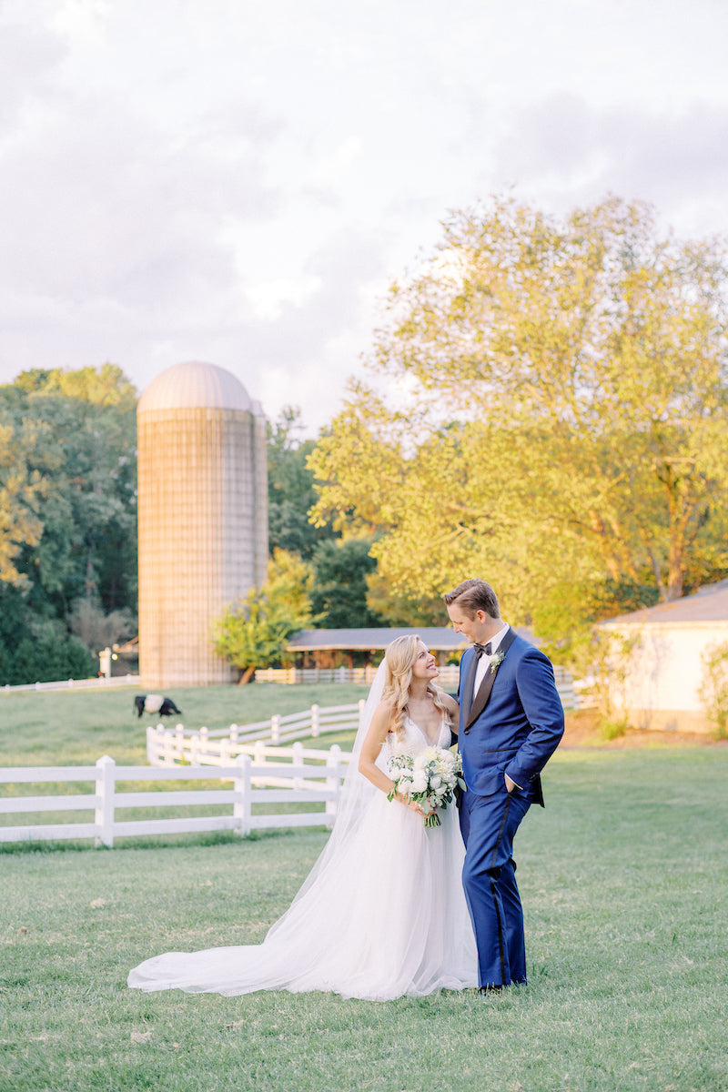 A bride and groom stand together on a grassy lawn. The bride wears a white gown and veil, holding a bouquet, while the groom wears a blue suit. A silo, white fence, and trees are in the background under soft sunlight.
