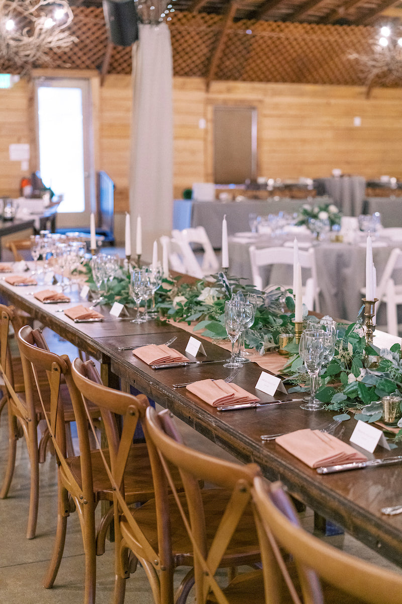 A long wooden table set for an event, decorated with greenery, candles, glassware, napkins, and place cards. Wooden chairs line the table in a rustic venue with wooden walls and other tables in the background.