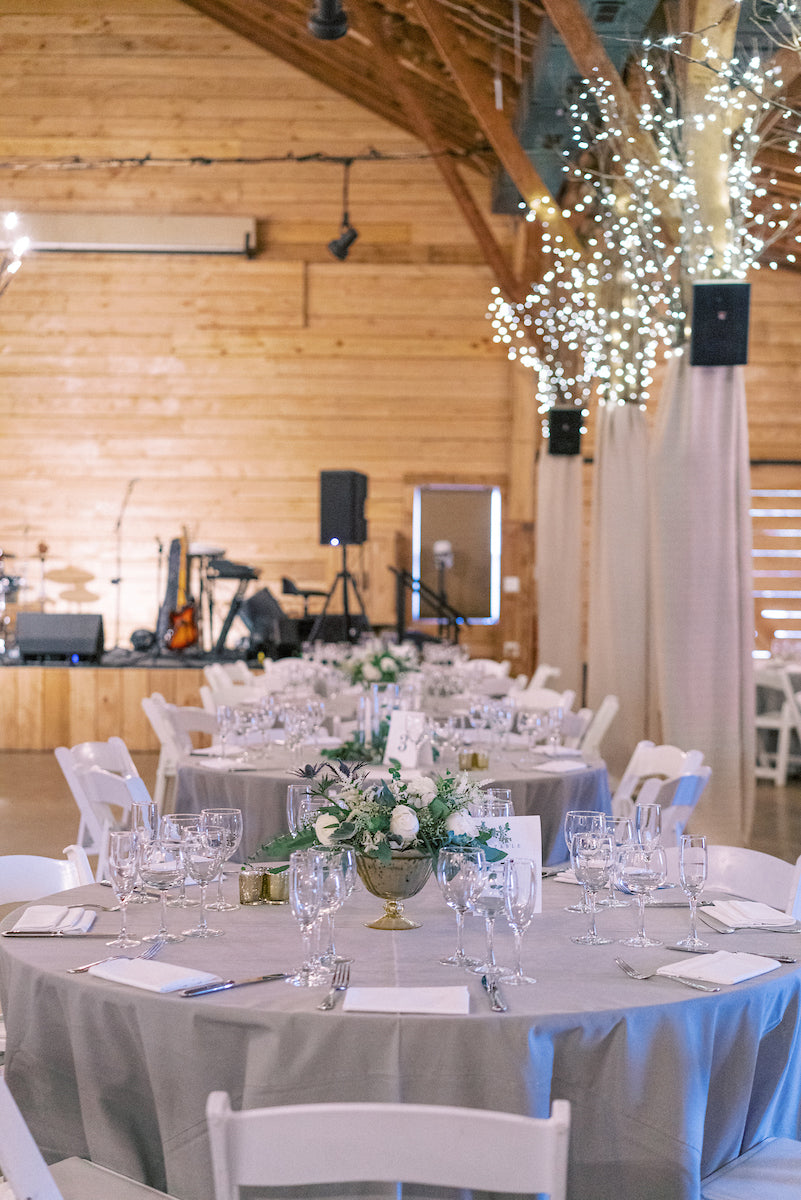 Round tables with gray tablecloths set for an event, decorated with floral centerpieces and glassware, in a wooden hall with twinkling string lights on beams and musical instruments visible on a stage in the background.