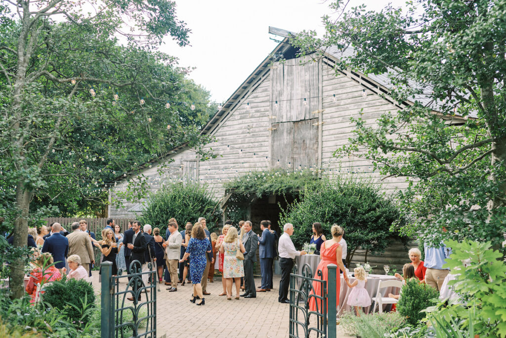 A group of people dressed in formal attire gather outside a rustic wooden barn surrounded by greenery, attending an outdoor event. Some guests sit and chat, while others stand and mingle under string lights.
