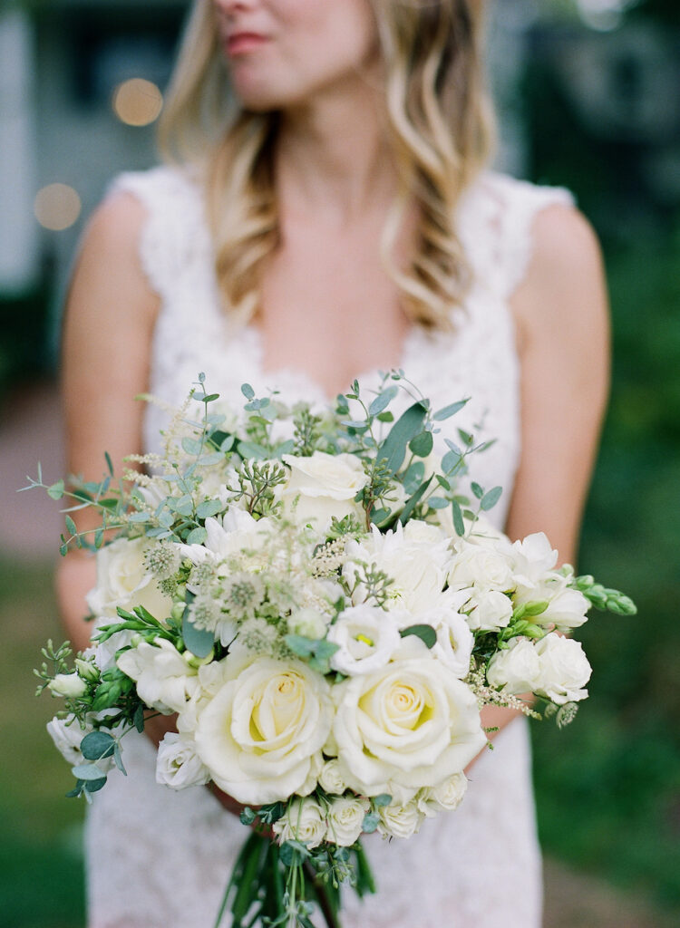 A woman in a white lace dress holds a bouquet of white and pale green flowers, including roses and greenery, outdoors. Her face is slightly out of focus and partially out of the frame.