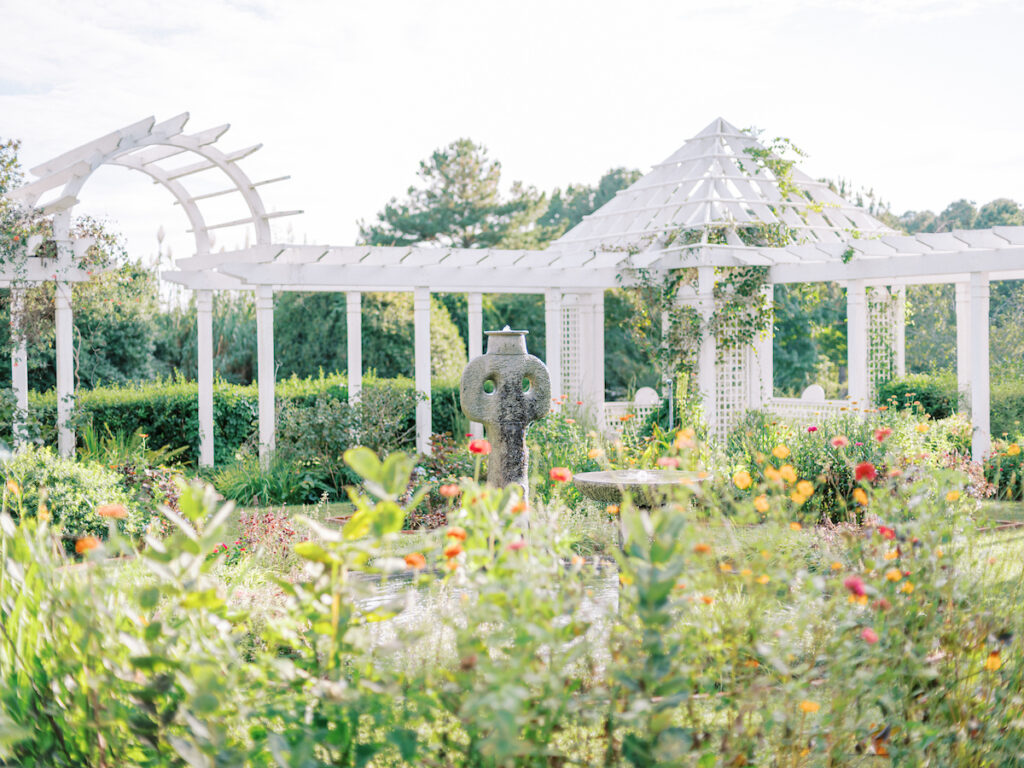 A lush garden with colorful flowers, a stone sculpture and fountain, and white pergolas surrounded by greenery under a bright, sunny sky.