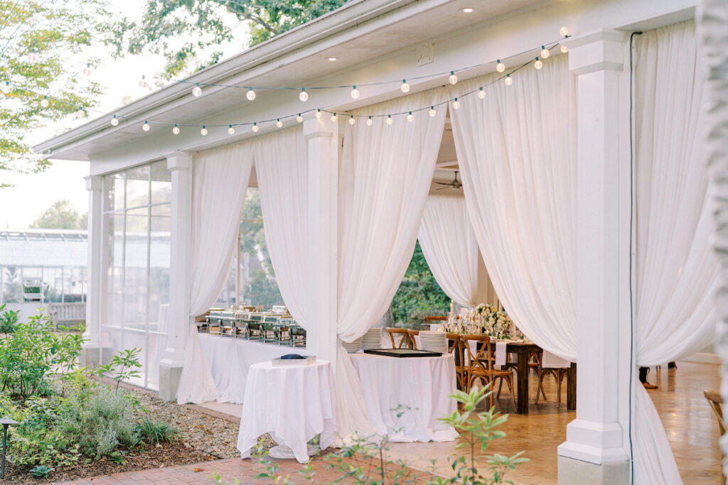 An elegant outdoor event space with white drapery, string lights, wooden chairs, and tables set for a gathering, surrounded by greenery and natural light.
