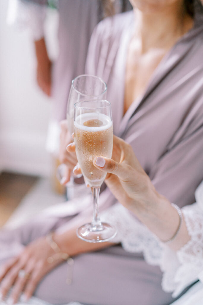 A woman in a lavender robe holds a glass of champagne, her nails painted light pink. She sits with another person in the background, both wearing robes with white lace trim. The scene feels celebratory and relaxed.