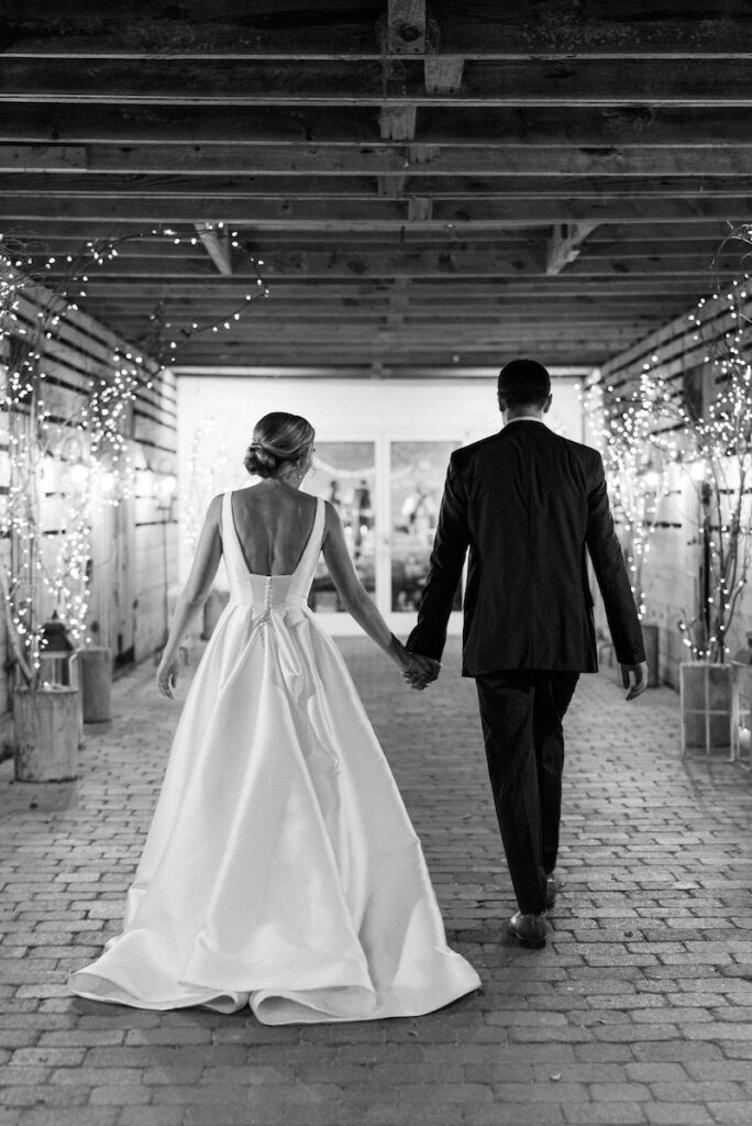 A bride and groom walk hand in hand under a wooden structure decorated with string lights. The bride wears a long, elegant gown, and the groom is in a suit. The scene is romantic and softly lit.
