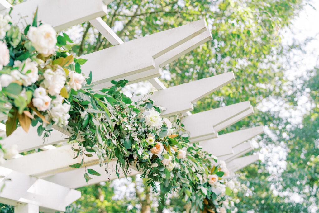 A white pergola decorated with lush green leaves and white flowers, set outdoors with sunlight filtering through green trees in the background.