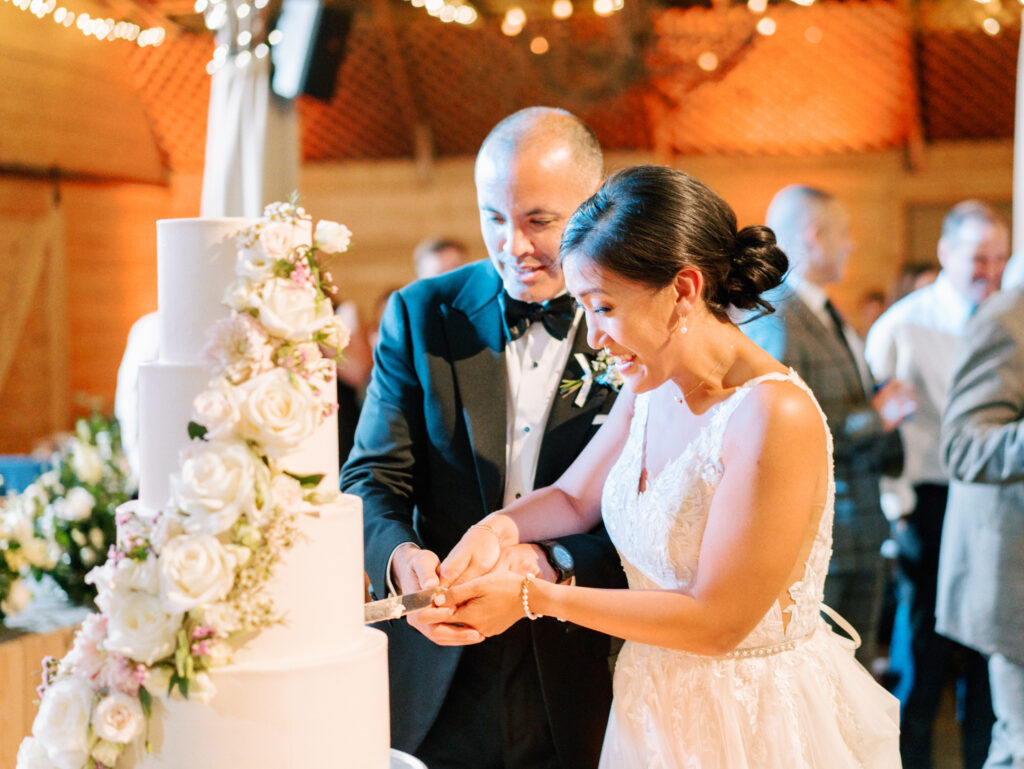 A bride and groom, smiling and dressed in formal attire, cut a tall white wedding cake decorated with white flowers at their reception, surrounded by guests in a warmly lit venue.