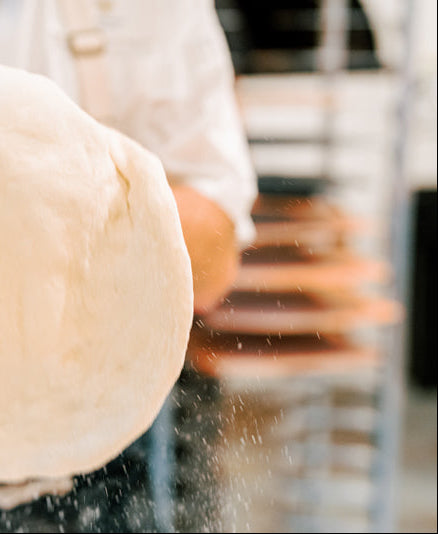 A person in a white shirt holding and tossing a large piece of dough, with flour particles visible in the air. The background is blurred, suggesting a kitchen or bakery setting.