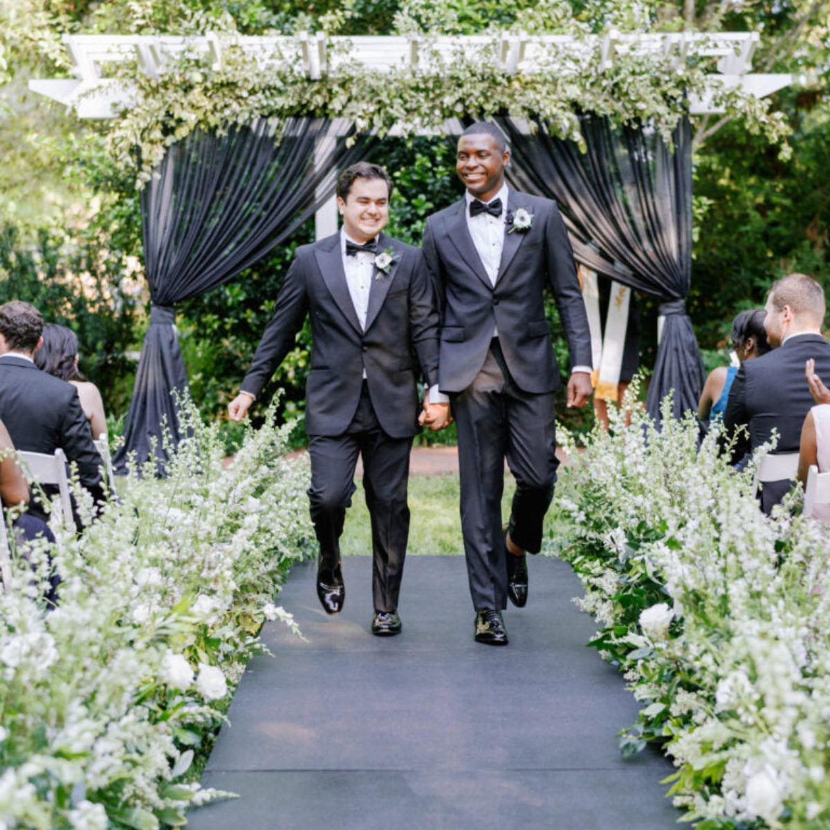 Two grooms in black tuxedos walk hand in hand down an outdoor aisle lined with white flowers and greenery, smiling as guests seated on either side watch and celebrate their wedding.