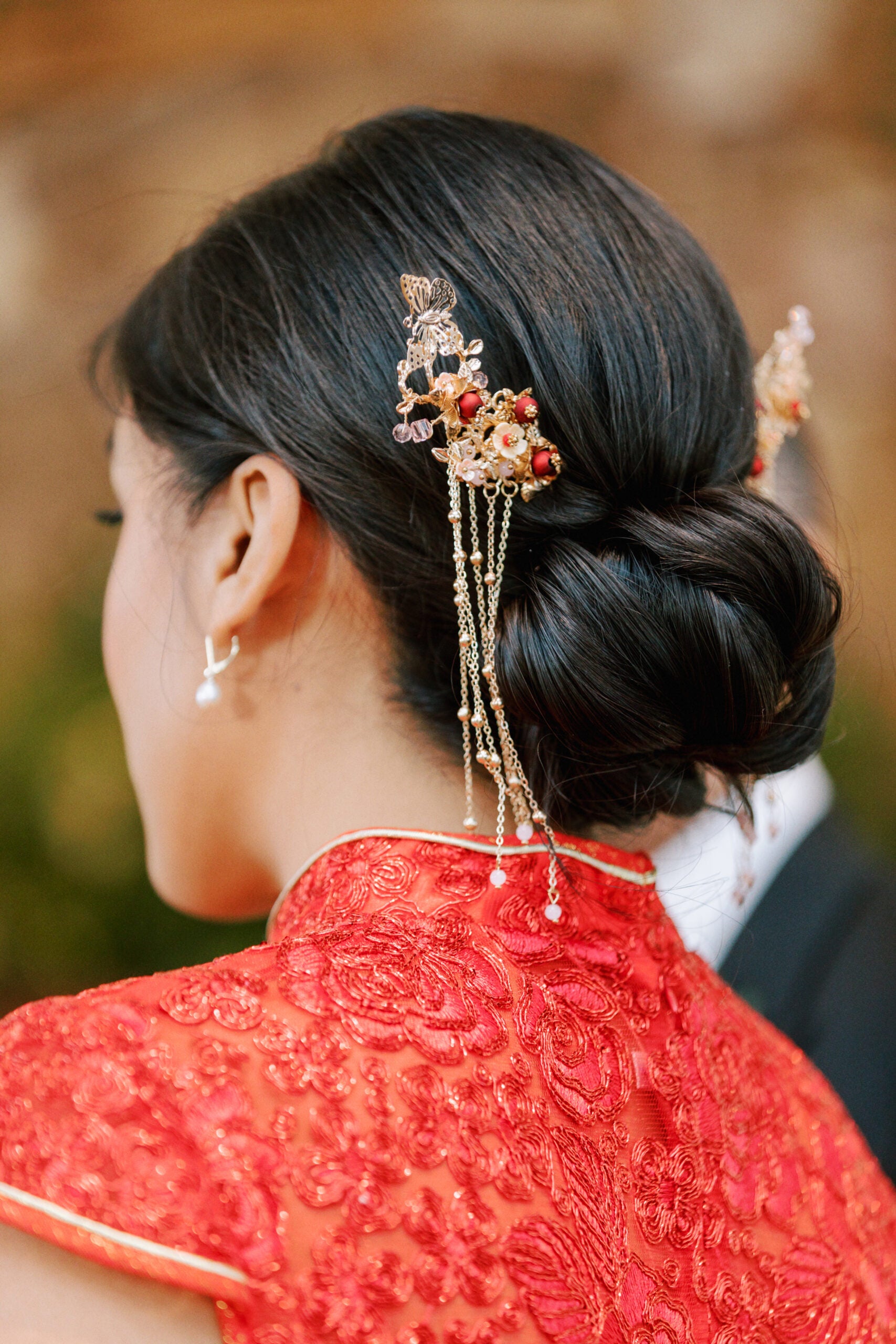 A woman wearing a traditional red dress with intricate gold embroidery has her dark hair styled in a low bun adorned with ornate gold hairpins and dangling chains, seen from the back and side.