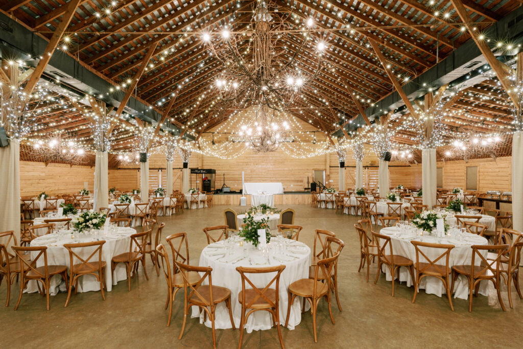A rustic barn venue decorated with string lights and chandeliers, featuring round tables with white tablecloths, wooden chairs, and floral centerpieces set for an event or wedding reception.
