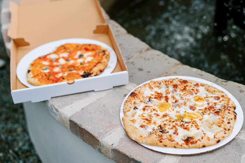 Two pizzas are displayed outdoors on a stone surface; one is on a white plate with cheese and yellow toppings, while the other, in an open box, has tomato sauce, cheese, and basil.